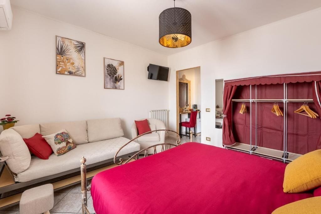 Un dormitorio con cama, sofá, sillón, radiador, televisor y ventana con cortinas.