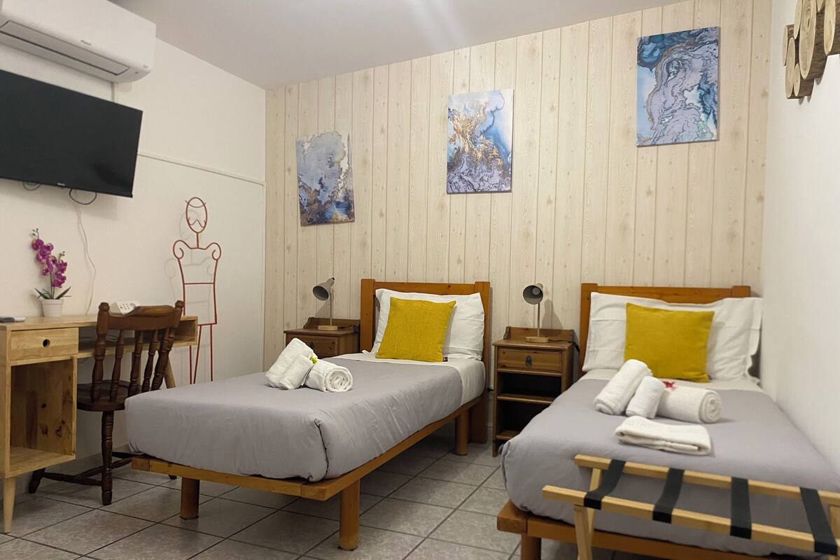 A hotel room with two single beds, a desk with a chair, a TV mounted on the wall, and wooden paneling.