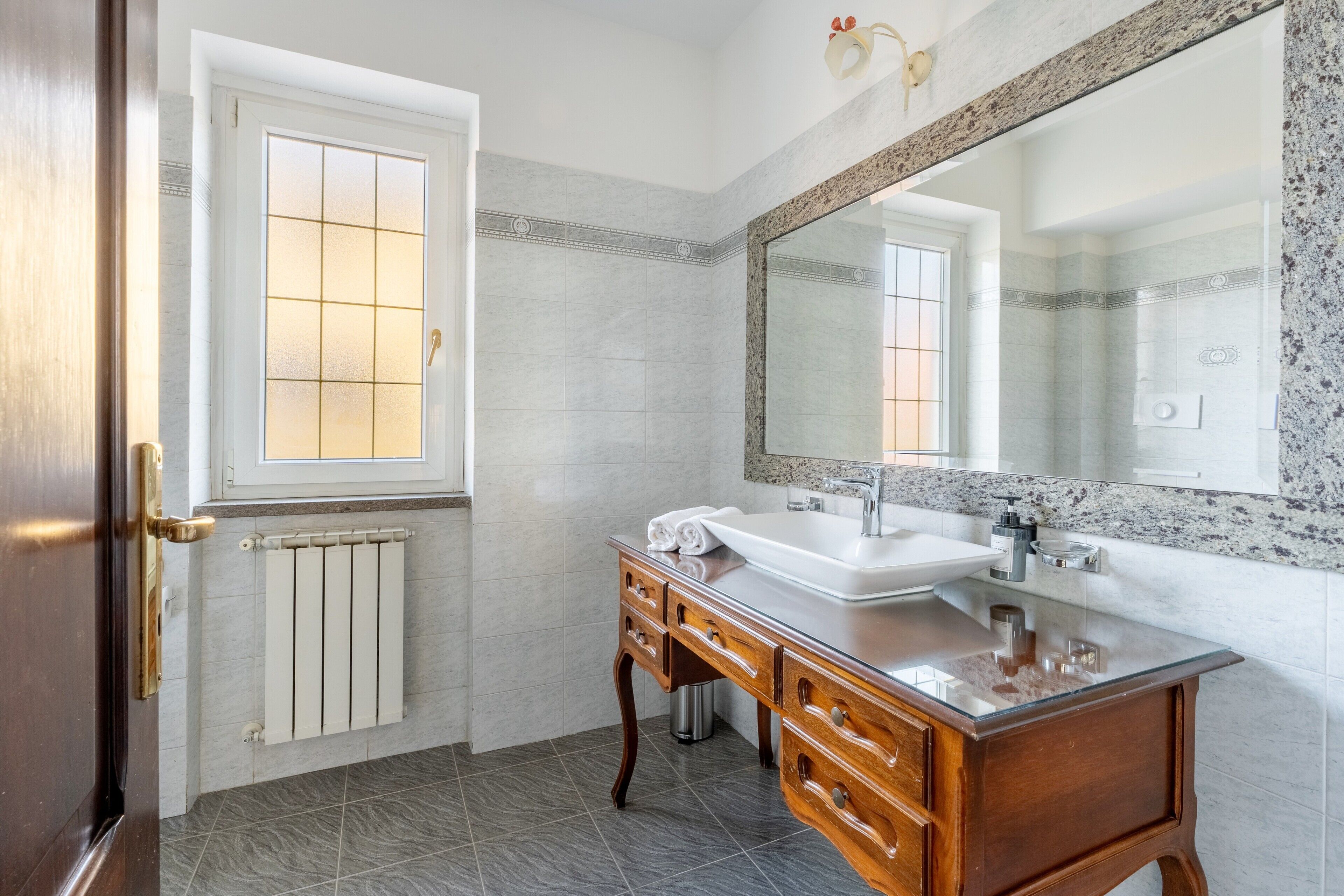 A bathroom with a wooden vanity, a large mirror, and a window with frosted glass.