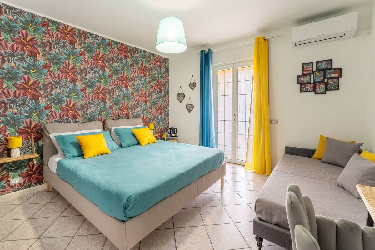 A bedroom with a bed, a sofa, a window with curtains, and a wall with a floral pattern.