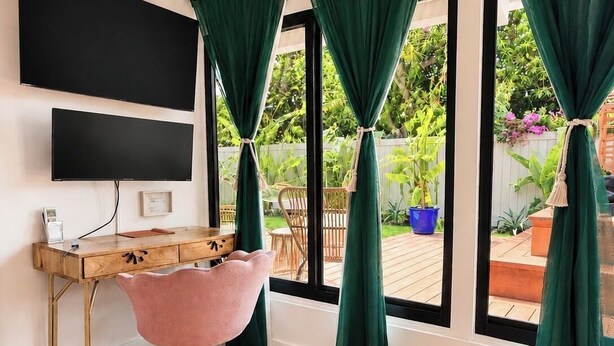 Una habitación con un escritorio de madera, una silla rosa, un televisor de pantalla plana y ventanas grandes con cortinas verdes.