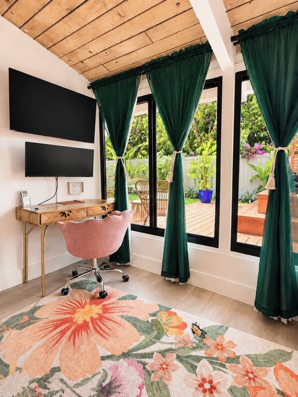 A room with a wooden desk, a pink chair, a flat-screen TV, and large windows with green curtains.