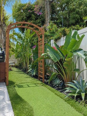 Un sendero en un jardín con césped artificial, una pérgola de madera y diversas plantas tropicales.