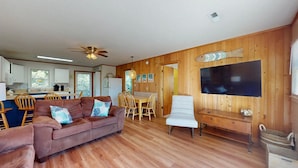 A cozy living room with a brown leather sofa, a dining table with chairs, a flat-screen TV mounted on the wall, and wooden paneling on the walls.