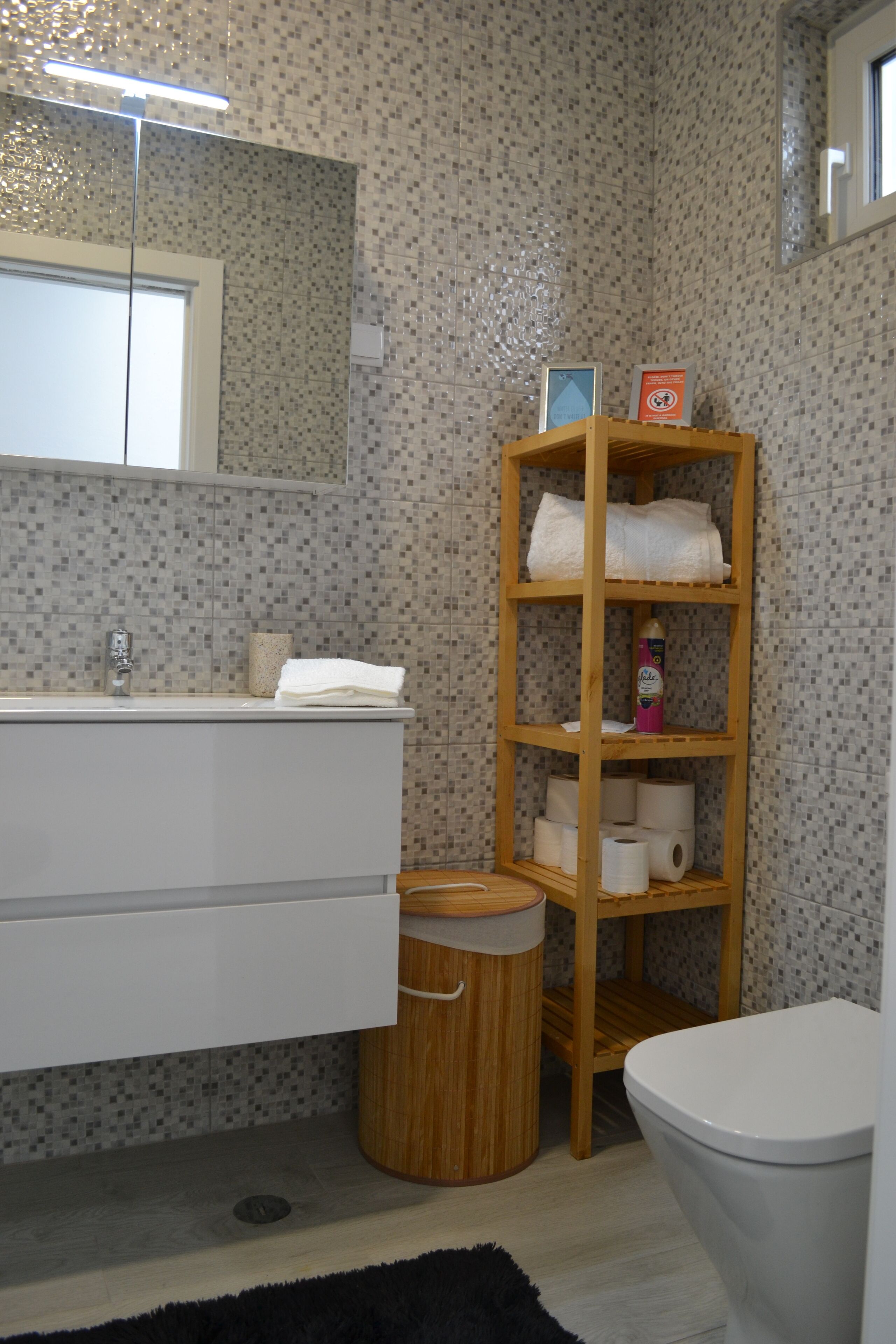 Un baño con un lavabo blanco, un espejo, un estante de madera con toallas, papel higiénico y un cubo de basura.