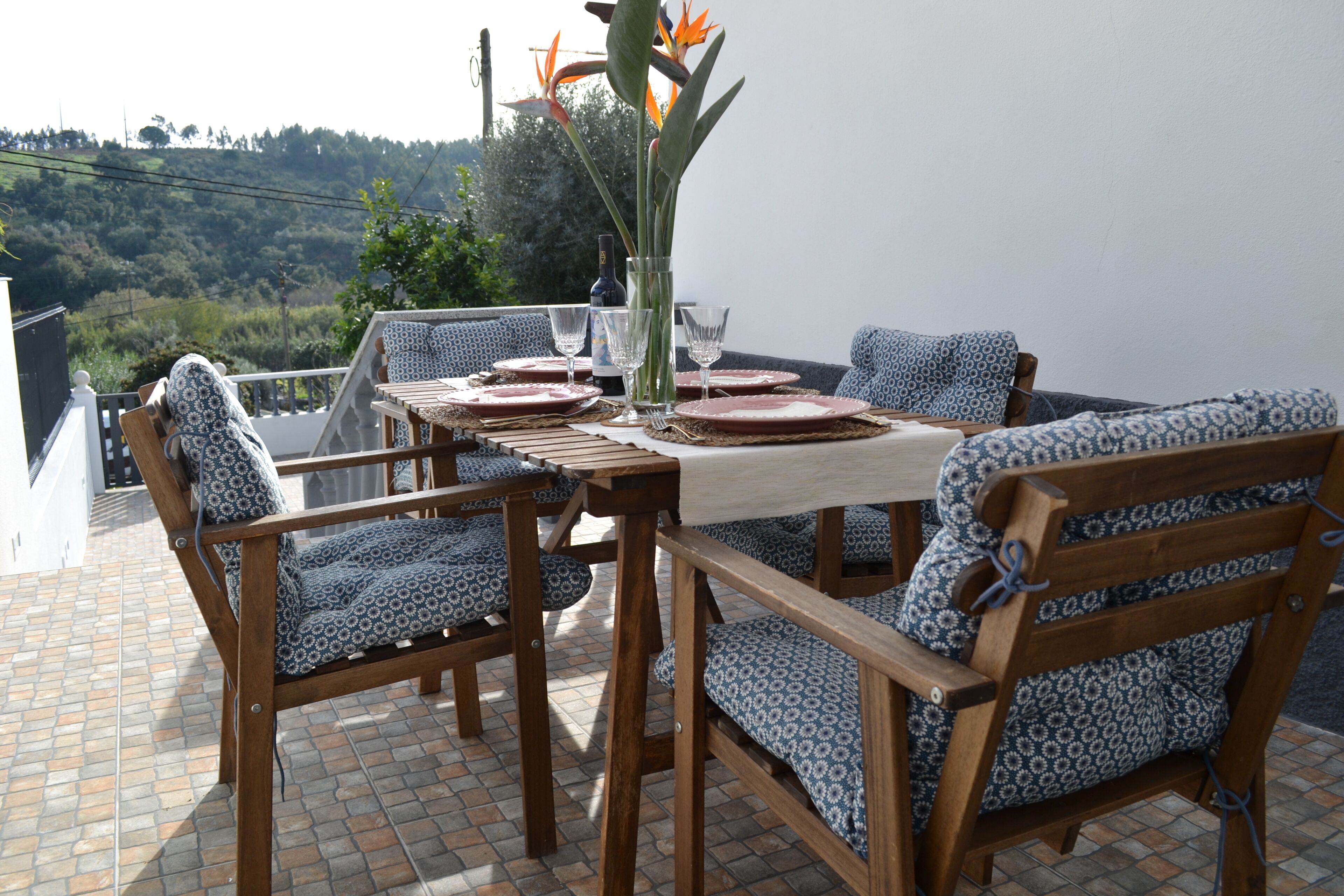 A terrace with a table set for four, surrounded by patterned chairs, a vase with flowers, and a view of a hillside.