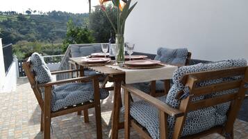 A terrace with a table set for four, surrounded by patterned chairs, a vase with flowers, and a view of a hillside.