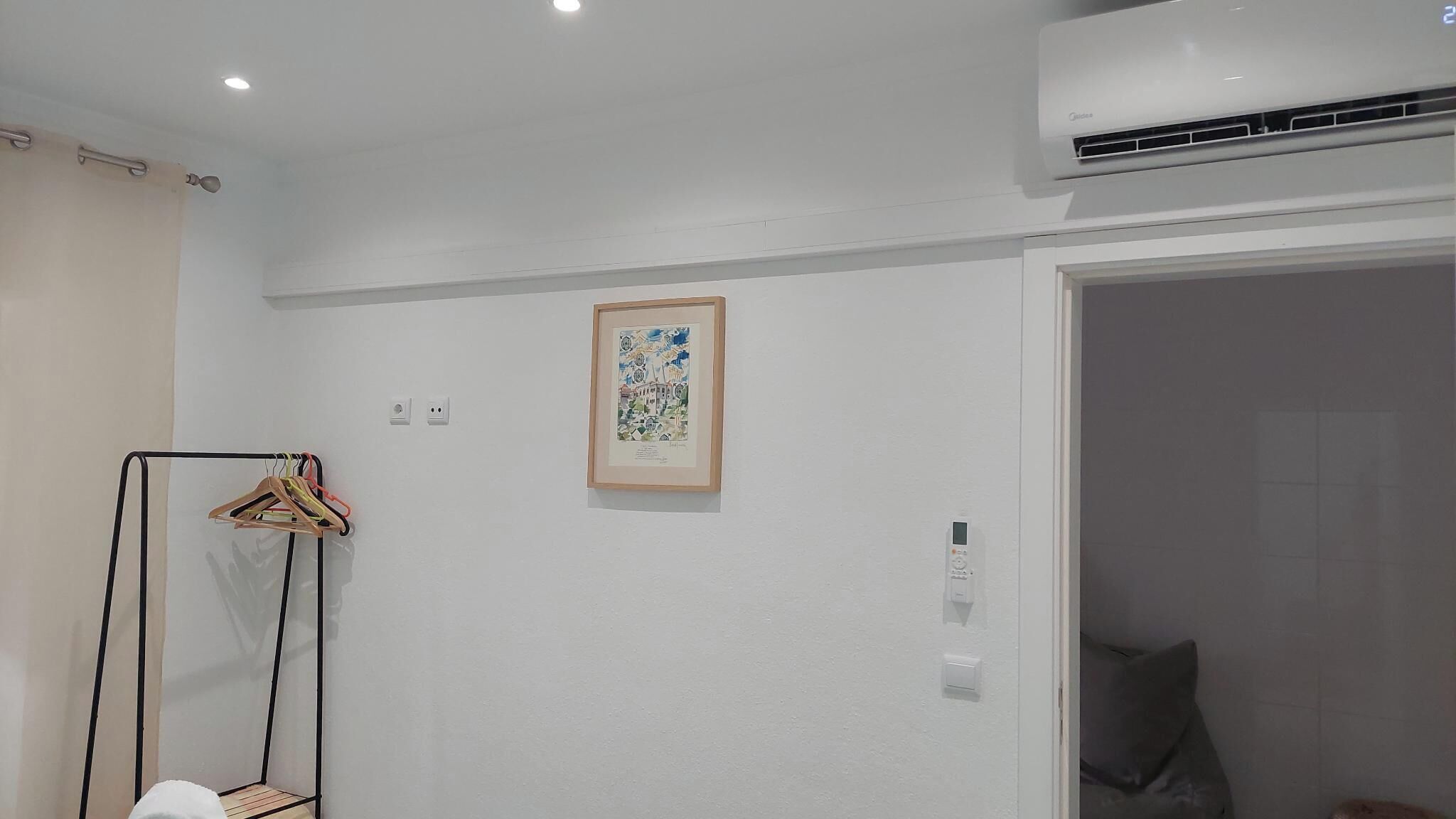A hotel room with a white wall, a framed picture, a coat rack, and an air conditioning unit.