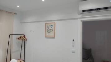 A hotel room with a white wall, a framed picture, a coat rack, and an air conditioning unit.