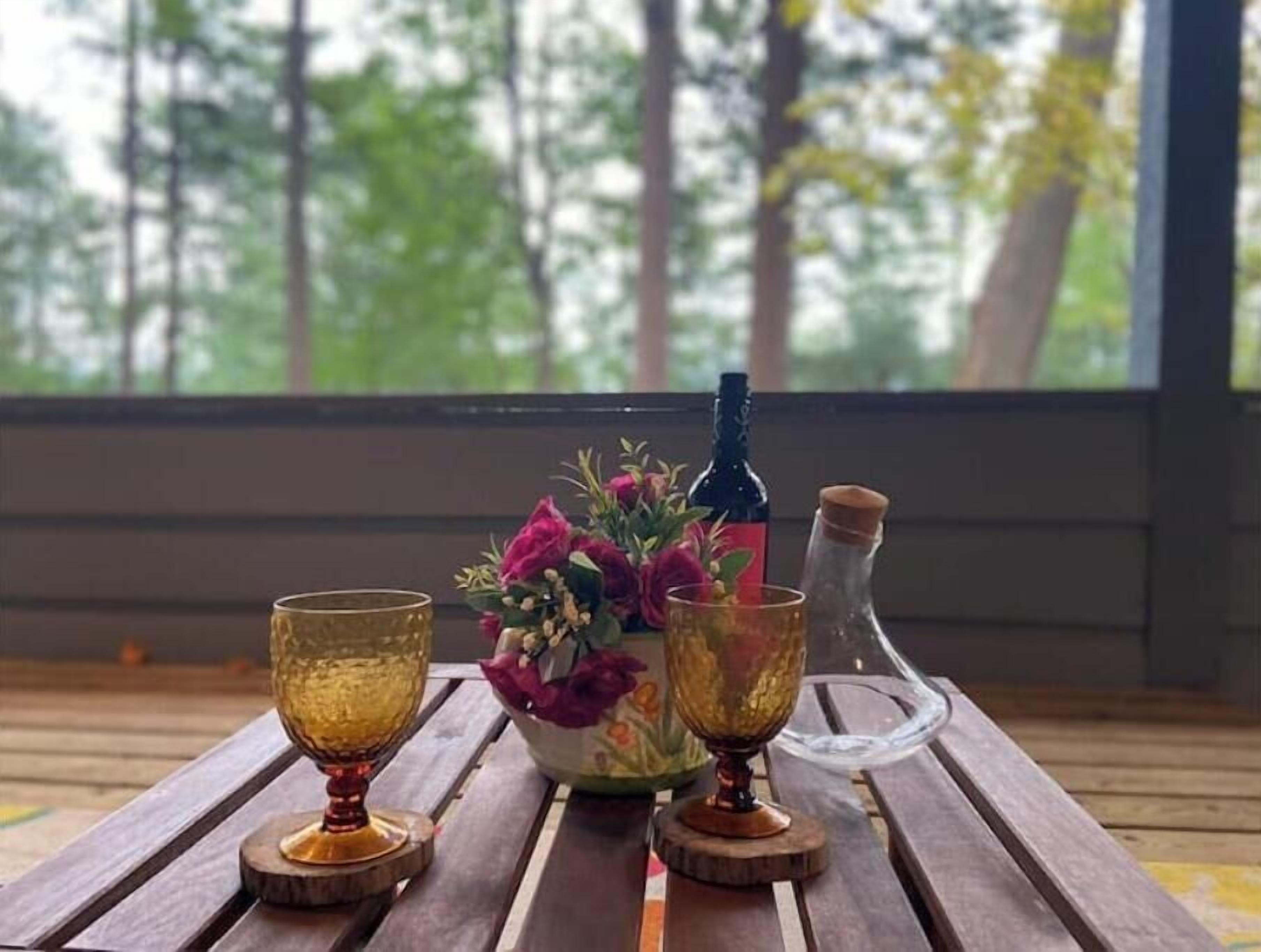 A wooden table with three glasses and a bottle on coasters, a vase with flowers, and a bottle of water on a porch with a view of trees.