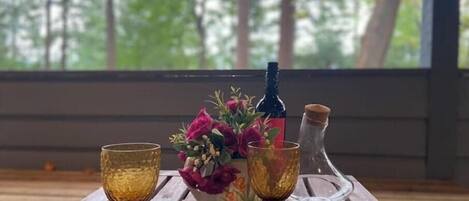A wooden table with three glasses and a bottle on coasters, a vase with flowers, and a bottle of water on a porch with a view of trees.