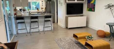 Una sala de estar moderna con un sofá, una mesa de centro y un televisor. Hay una zona de cocina con un refrigerador y una mesa de comedor.