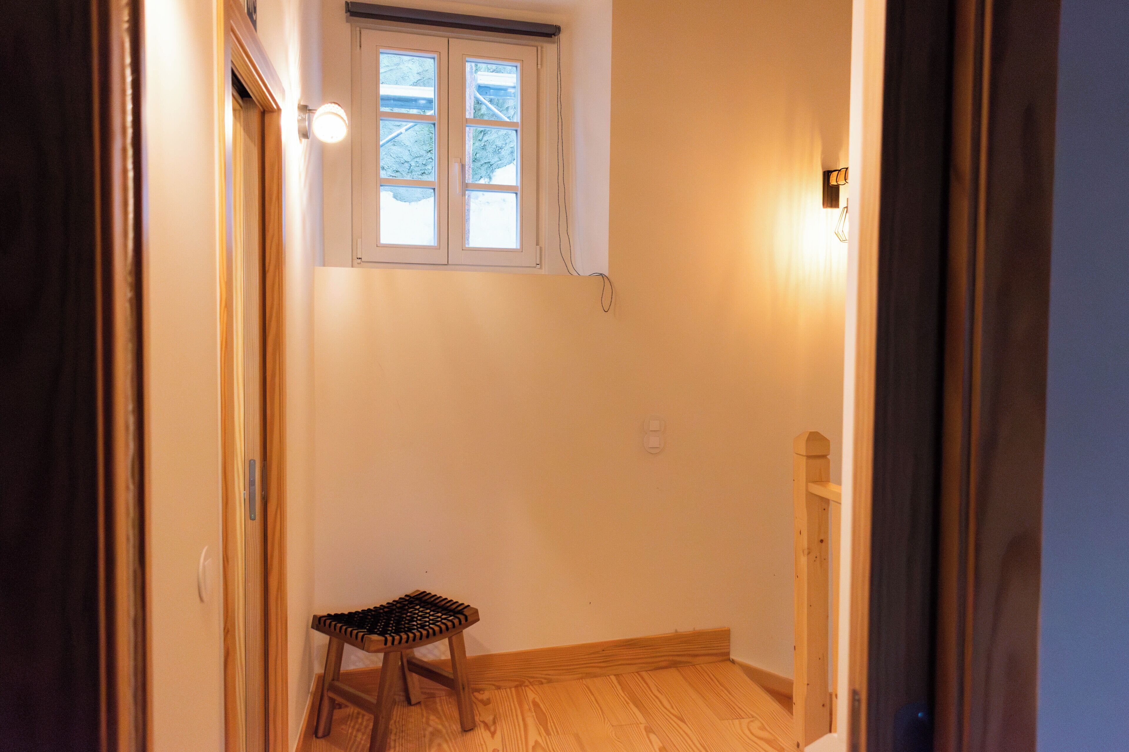A narrow hallway with a small wooden stool, a window with a view of trees, and a light fixture on the wall.