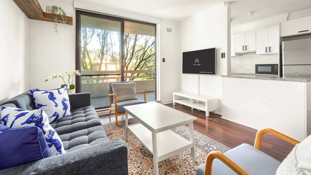A modern living room with a sofa, coffee table, and a TV. There's a kitchen with white cabinets and a microwave.