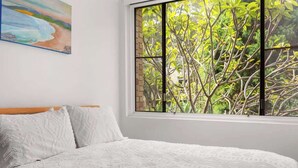 A bedroom with a large window, a bed with patterned bedding, and a painting on the wall.