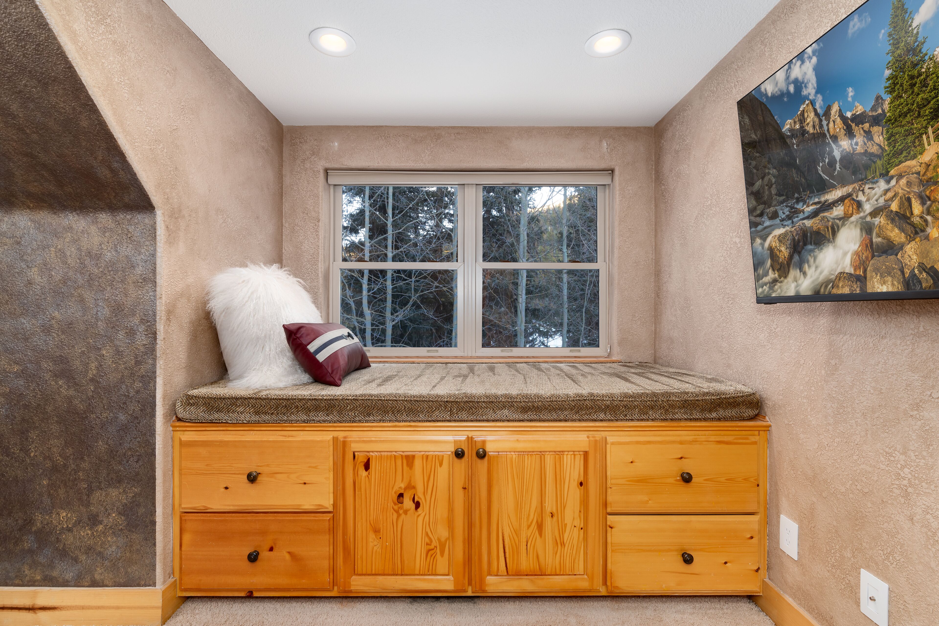 A cozy nook with a cushioned bench, a window with a view of trees, and a painting of a mountain landscape.