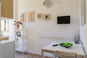 Ein moderner, minimalistischer Raum mit einem weißen Tisch, einem Stuhl, einem Regal mit einer Pflanze, einem Fernseher und Kunstwerken an der Wand.