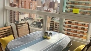 A dining area with a table and chairs, a view of a cityscape through large windows, and a balcony with a railing. - Gemelos 22, Benidorm-Alicante (Benidorm)
