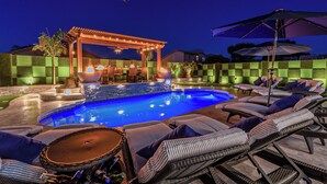 Ein Pool mit Liegestühlen und Sonnenschirmen, umgeben von einer Schachbrett-Mauer und einer Pergola mit Beleuchtung.