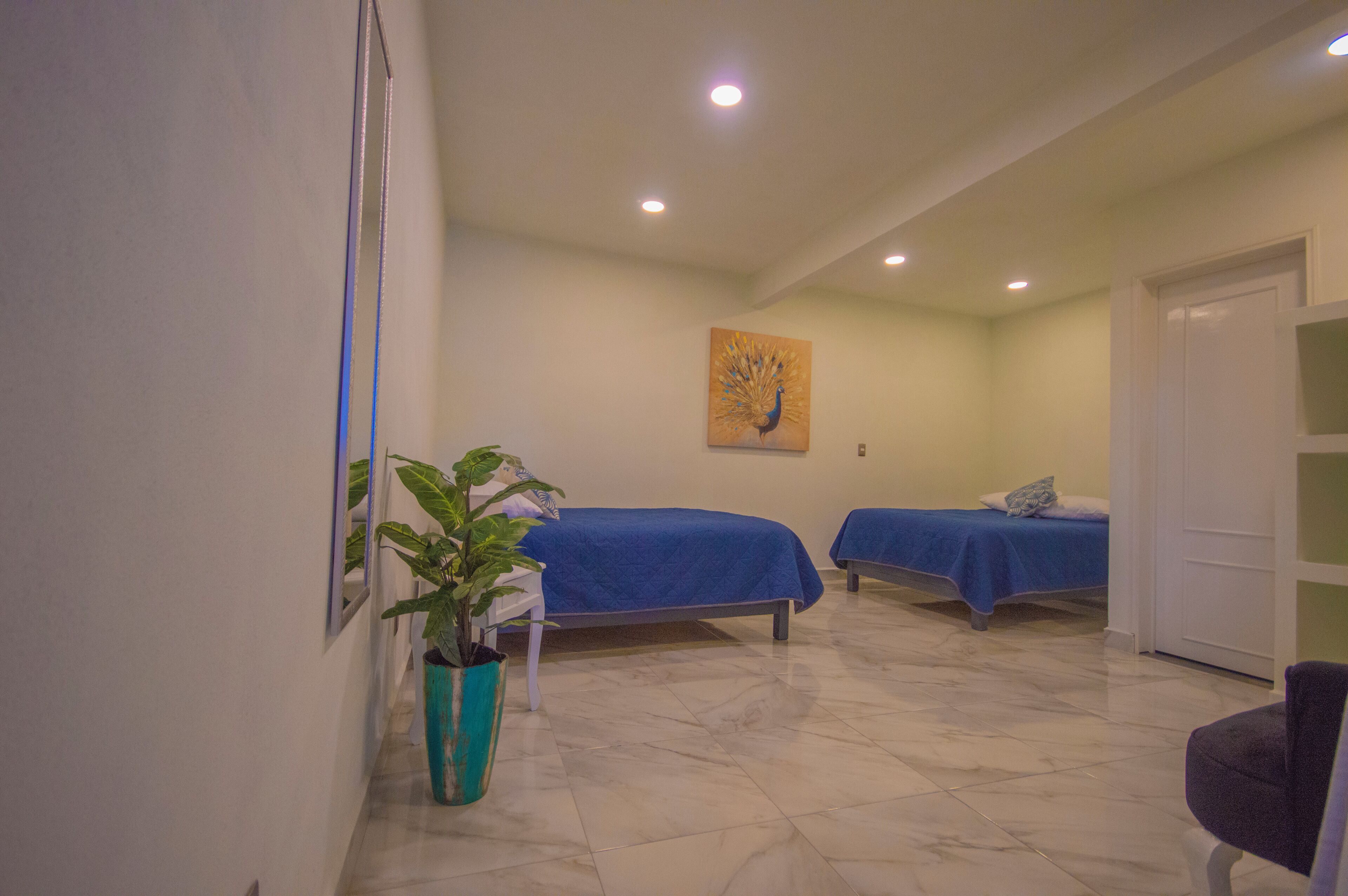 A room with two beds, a potted plant, and a painting on the wall.