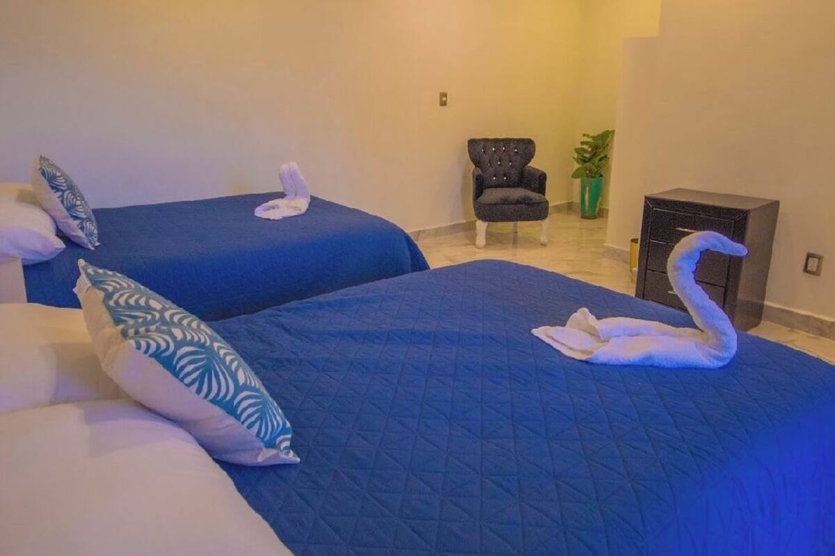 Ein Hotelzimmer mit zwei Betten, jeweils mit blauer Steppdecke und weißen Kissen. In der Ecke steht ein Sessel und ein Blumentopf.