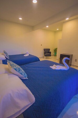 Een hotelkamer met twee bedden, elk met een blauw geborduurd sprei en witte kussens. In de hoek staat een stoel en een potplant.