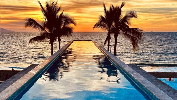 Una piscina infinita con vista al océano y palmeras al atardecer.