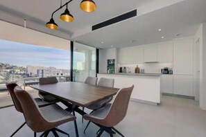 Eine moderne Küche mit weißen Schränken, eine Essecke mit Holztisch und Stühlen sowie große Fenster mit Blick auf die Stadt.