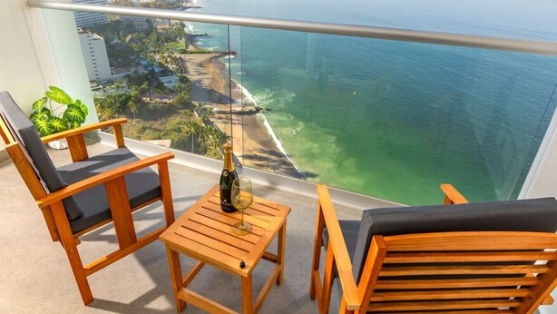 A balcony with wooden furniture, a bottle of champagne, and a glass on a small table, overlooking a coastal city and ocean.