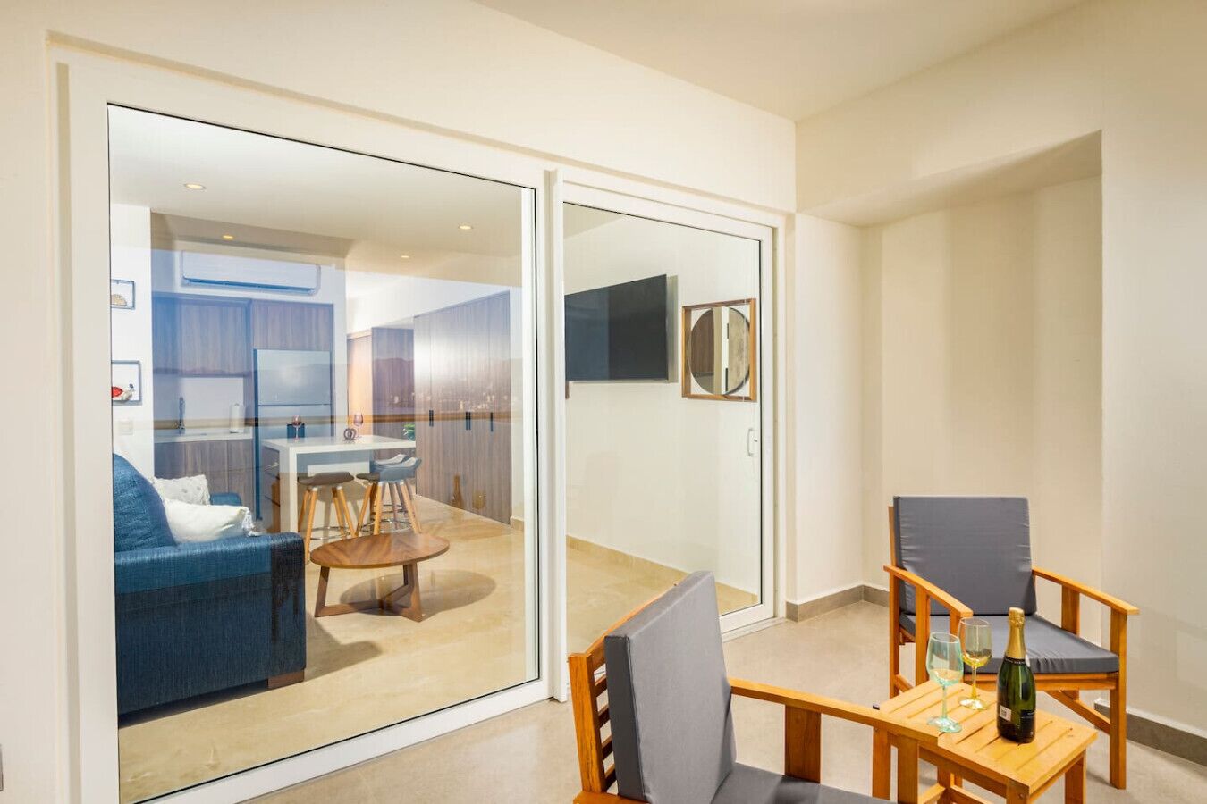 A modern living room with a glass door leading to a kitchen and dining area. There is a wooden table with two chairs and a bottle of wine on it.