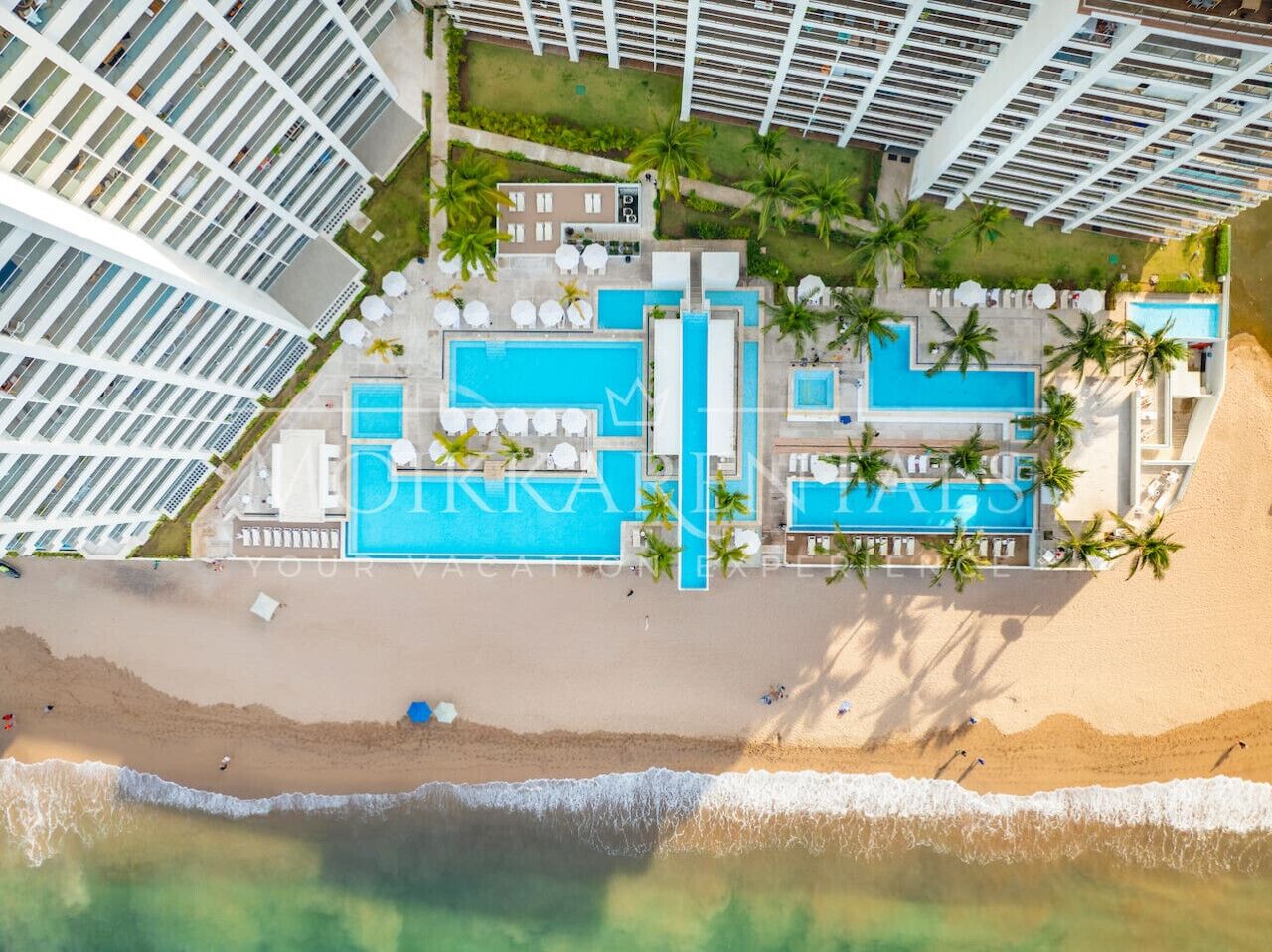 Vista aérea de un complejo turístico con múltiples piscinas, palmeras y acceso a la playa.