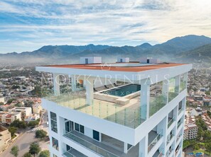 Un edificio moderno con piscina en la azotea y barandillas de cristal, que ofrece vistas a una ciudad y montañas al fondo.