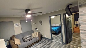 Ein kleiner Wohnraum mit grauem Sofa, Deckenventilator, Bett mit blauer Bettdecke und Spiegel, der den Raum reflektiert.