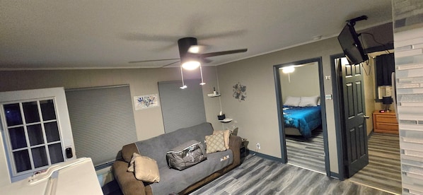 Ein kleiner Wohnraum mit grauem Sofa, Deckenventilator, Bett mit blauer Bettdecke und Spiegel, der den Raum reflektiert.