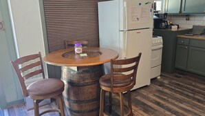 Una cocina con una mesa redonda de madera, dos sillas, un refrigerador blanco, una estufa y un cubo de basura.