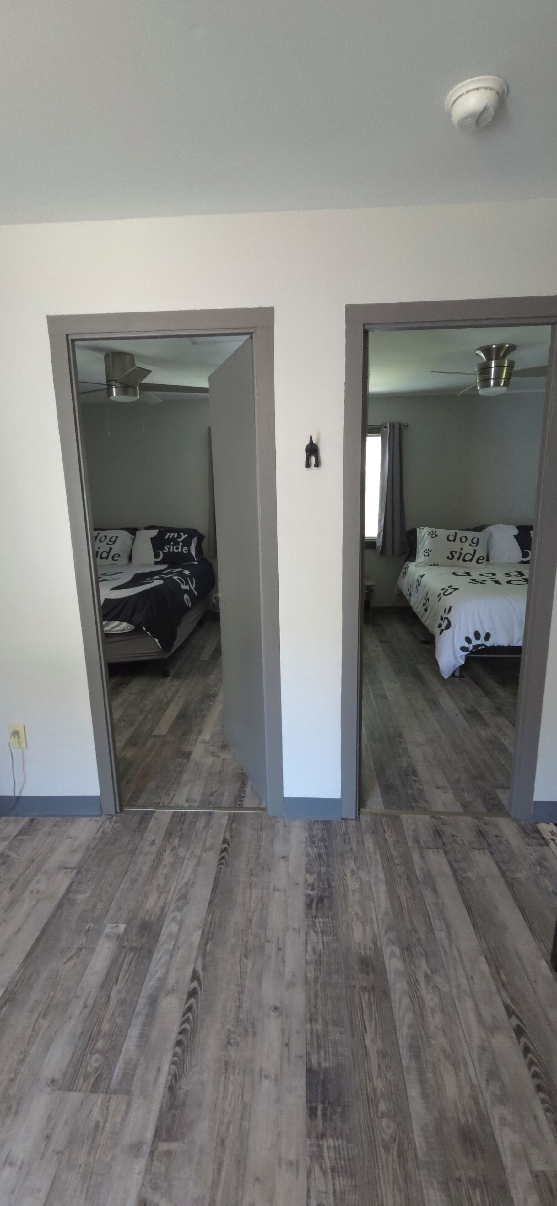 Une pièce avec un plancher en bois, un lit recouvert d’une couvre-lit à motifs noirs et blancs, un ventilateur de plafond et un interrupteur visible sur le mur.