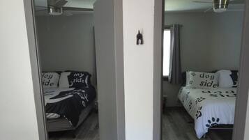 Une pièce avec un plancher en bois, un lit recouvert d’une couvre-lit à motifs noirs et blancs, un ventilateur de plafond et un interrupteur visible sur le mur.