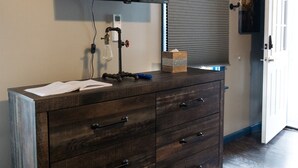 Un baño con tocador de madera, televisor montado y ventana con persianas.