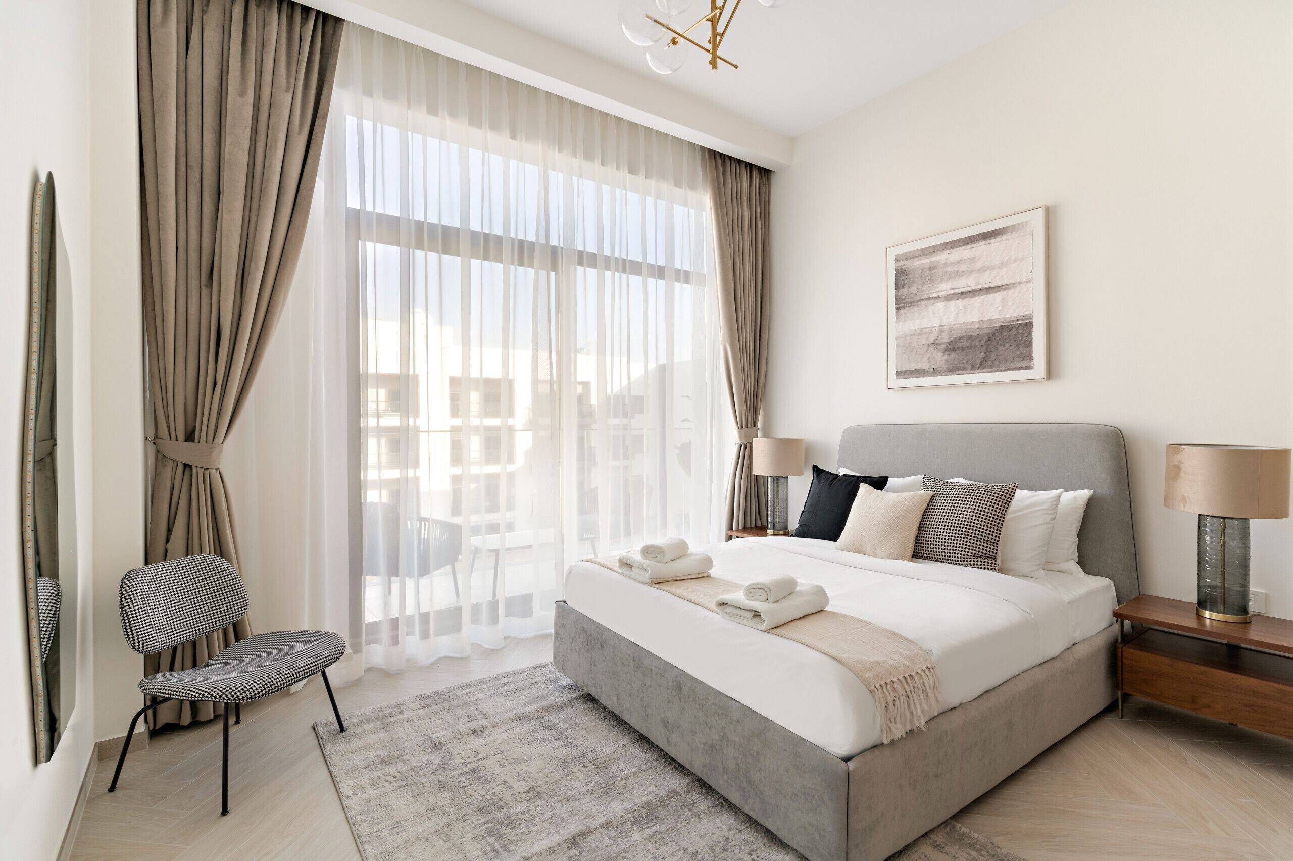 A modern bedroom with a large bed, a chair, a bedside table with lamps, and sheer curtains.