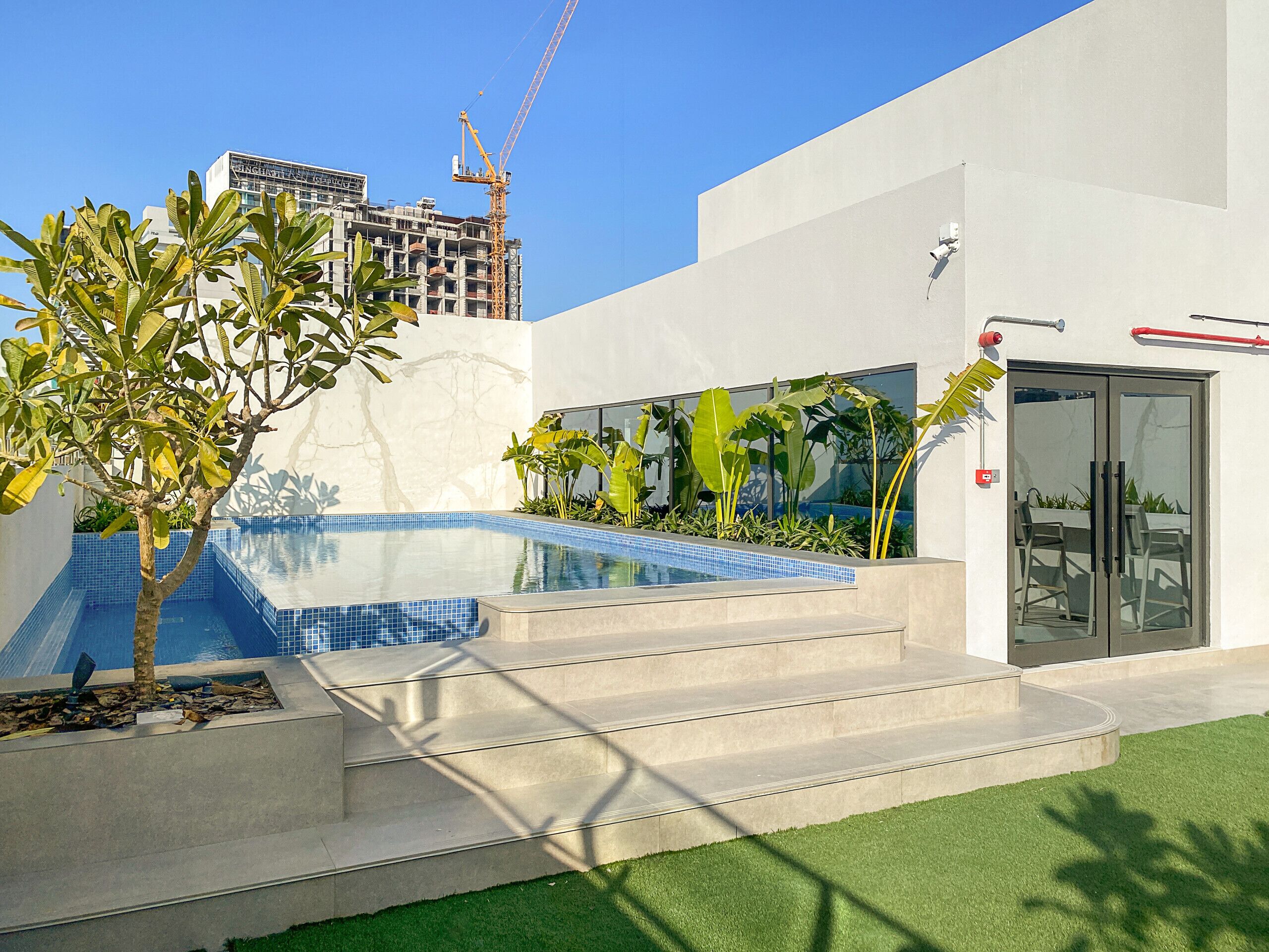 Zona de piscina exterior moderna con escalones que conducen a una puerta de cristal, rodeada de vegetación y un cielo azul despejado.