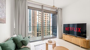 Ein modernes Wohnzimmer mit großem Fernseher, einem grünen Sofa und Blick auf hohe Gebäude durch raumhohe Fenster.