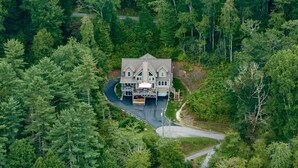 Une grande maison à deux étages, entourée d’une forêt dense, avec une allée menant à une entrée fermée par un portail.
