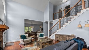 Un salon spacieux, doté d’un grand canapé, d’une table basse et de deux fauteuils. Un escalier avec une rampe en bois mène à l’étage supérieur.