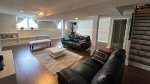 A spacious living room with a black leather sofa, a white coffee table, and a patterned rug. There is a bookshelf with books and decorative items, and a window with curtains.