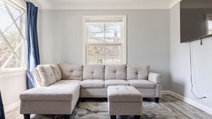A living room with a sectional sofa, ottoman, and a large window with curtains. - Cozy Euclid Home Near Lakefront (Euclid)