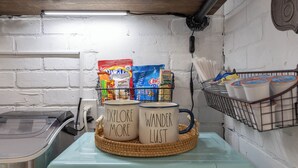 A kitchenette with a retro-style appliance, a basket with two mugs, and a shelf with food items and utensils. - Gypsy Essence: Free Parking | Steps to Downtown (Eureka Springs)