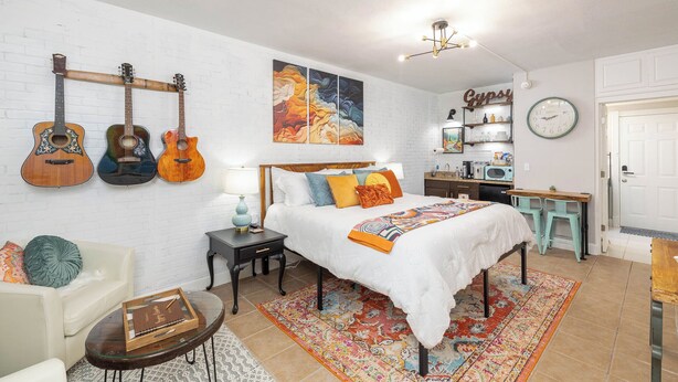 A bedroom with a bed, a nightstand, a chair, a desk, and a wall with guitars and a painting. - Gypsy Essence: Free Parking | Steps to Downtown (Eureka Springs)