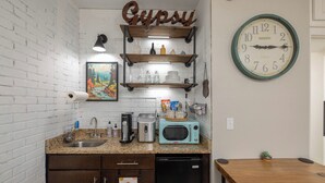A kitchen with a brick wall, a wooden shelf with kitchenware, a microwave, and a clock. - Gypsy Essence: Free Parking | Steps to Downtown (Eureka Springs)