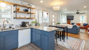 A modern kitchen with blue cabinets, a dishwasher, and a granite countertop. There is a dining area with a table and chairs, and a living room with a sofa and a TV.