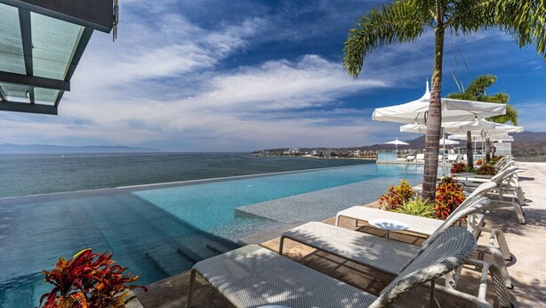 Une piscine à débordement offrant une vue sur la mer et une île lointaine. Des chaises longues et des parasols sont disposés le long du bord de la piscine.